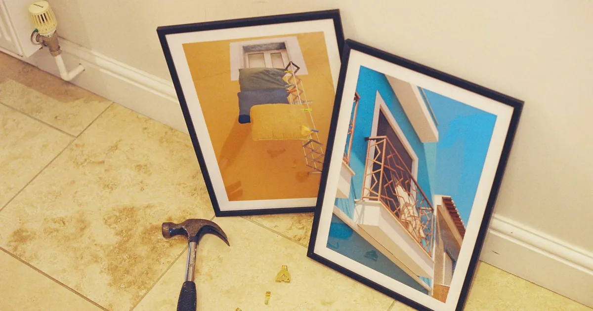 Two framed prints leaning against a wall next to a hammer and picture hooks on a tiled floor
